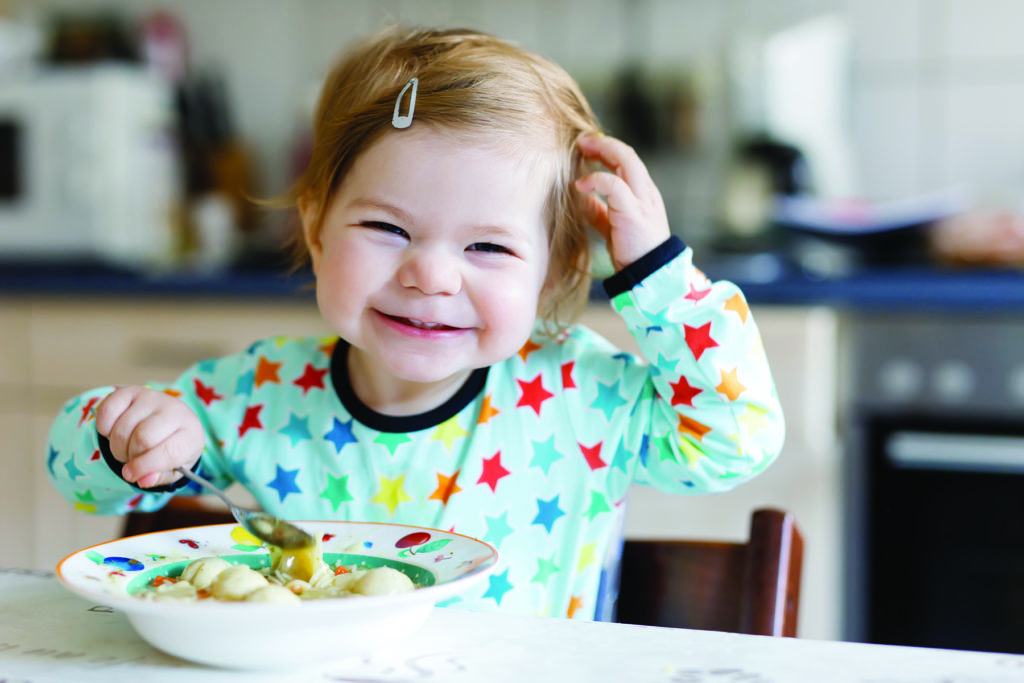 Adorable baby girl eating from spoon vegetable noodle soup. food, child, feeding and development concept. Cute toddler, daughter with spoon sitting in highchair and learning to eat by itself