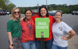 Group of 4 female Food Bank team members gather around a green sign reading Trail Starts