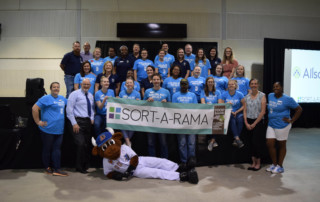 Group of 40 Food Bank team members in matching blue t-shirts pose with Sort-A-Rama Sign. Wool E Bull the baseball mascot lays in front of group. 