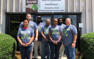 Group of 7 Sandhills branch staff members gather in matching food bank t-shirts in front of the Sandhills Branch building.