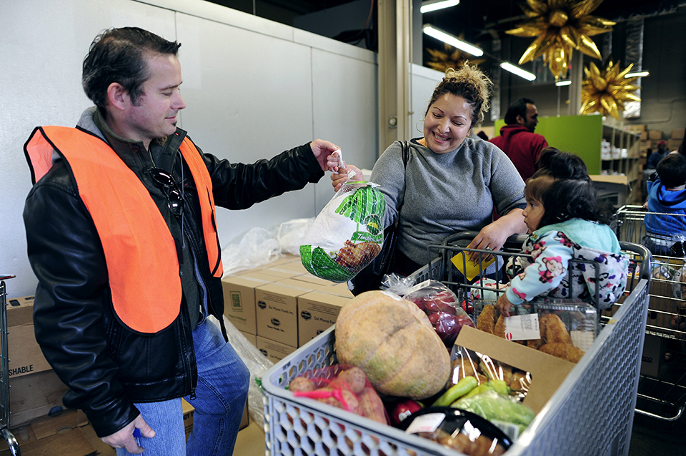 A volunteer hands a turkey to a smiling woman behind a packed grocery cart with a child in the front seat.