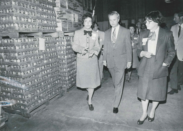 Black and White; 3 People walk through new warehouse surrounded by stacked boxes in 1983