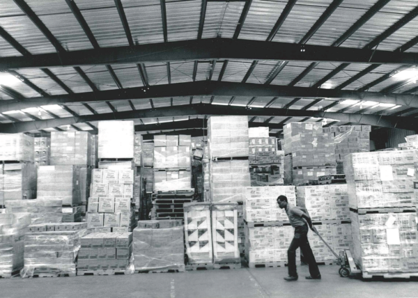 Black and white photo; Man pulling pallet jack stacked with boxes through a warehouse in 1990