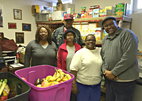 5 individuals gathered in a small pantry surrounded by bins of produce and shelved staple items