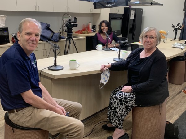 Peter Werbicki and Barbara Oates smile at the camera in a kitchen with podcast microphones