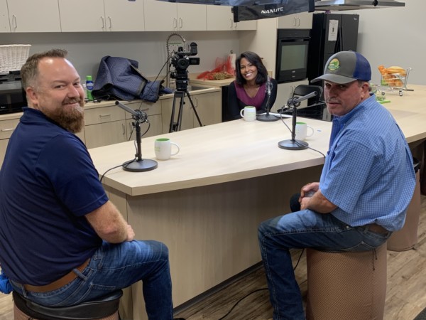 Carter Crain and Bill Hering smile at the camera in a kitchen with podcast microphones