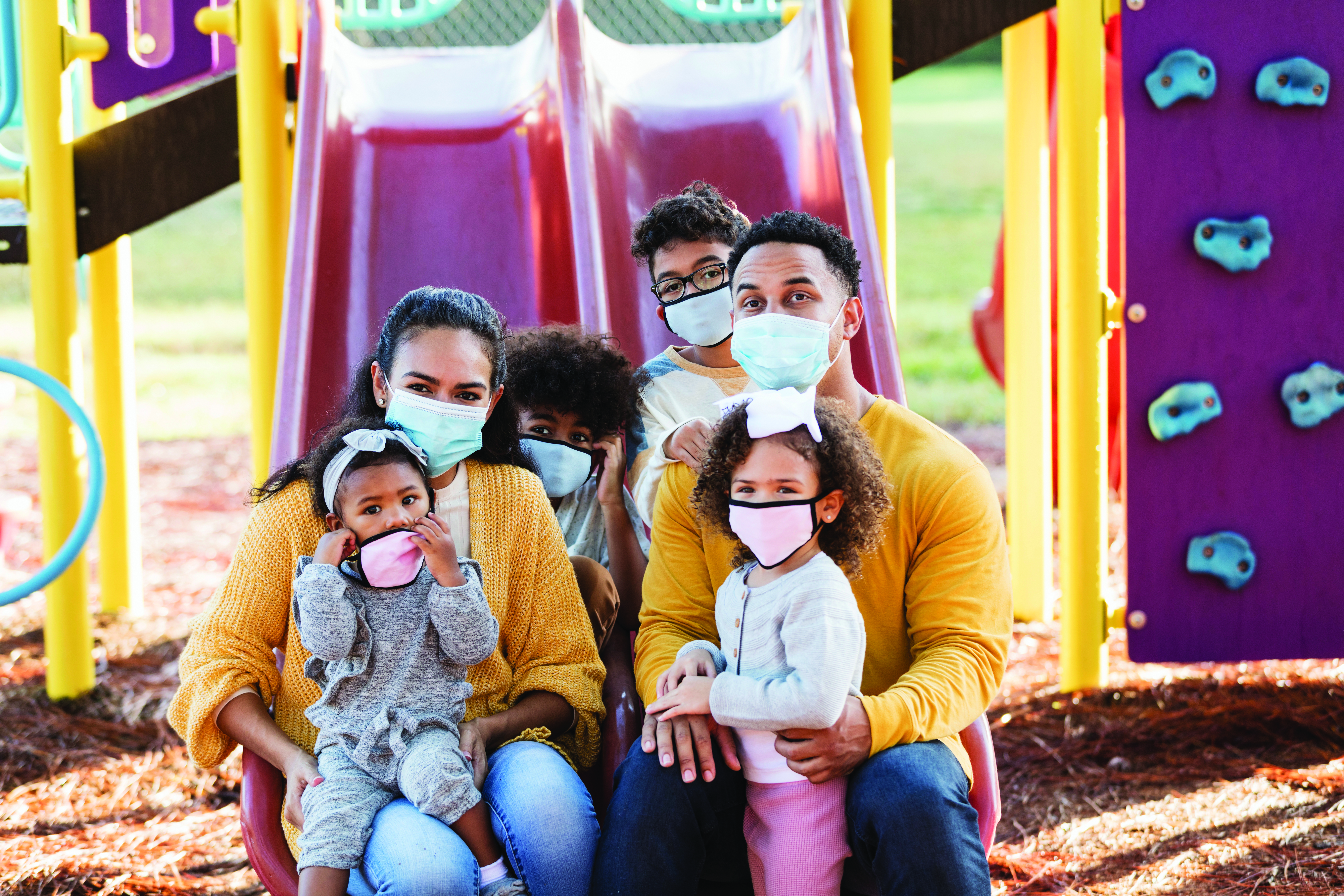 Family of 6 sits together at a playground in front of two red slides each wearing a mask