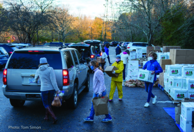 Masked individuals carry boxes and bags of food from stacks on the right to a line of cars waiting with trunks open on the left