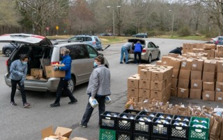 Masked volunteers load a van with boxes from a stack to the right at a drive through distribution