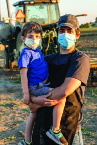 Man wearing a mask stands in front of a tractor holding a child also wearing a mask
