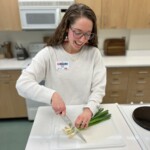 Rachel Ezzell with long hair and glasses wearing a light colored sweater is cutting vegetables on a cutting board.