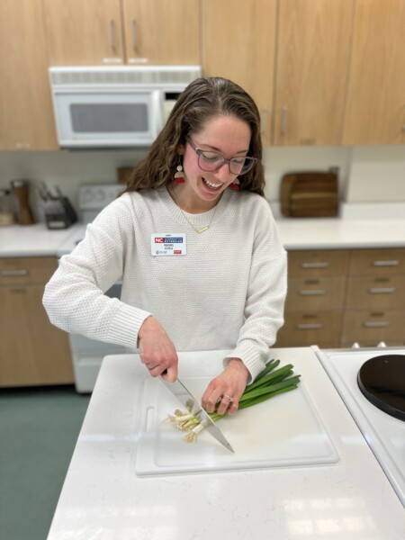 Rachel Ezzell with long hair and glasses wearing a light colored sweater is cutting vegetables on a cutting board.